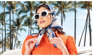 Uma jovem com um vestido laranja vibrante está parada ao lado de um carro clássico amarelo e branco sob palmeiras. Ela usa óculos de sol brancos grand
